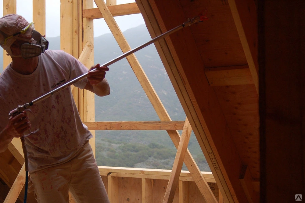 Worker applying spray foam insulation to attic ceiling with a spray gun while wearing protective gear.