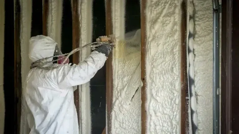 Worker in protective gear spraying foam insulation in long island between wall studs .