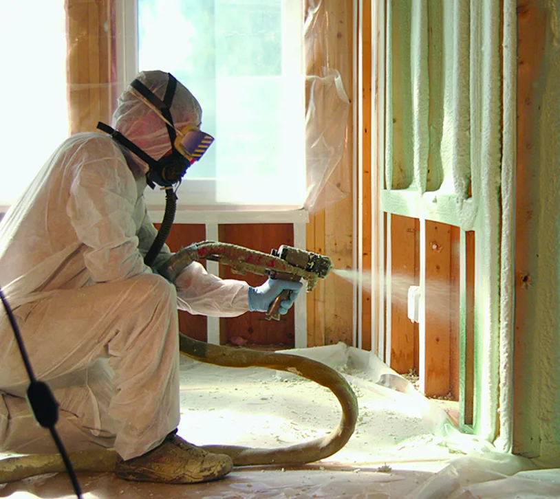Technician in protective gear applying spray foam insulation to wall framing in a residential interior.