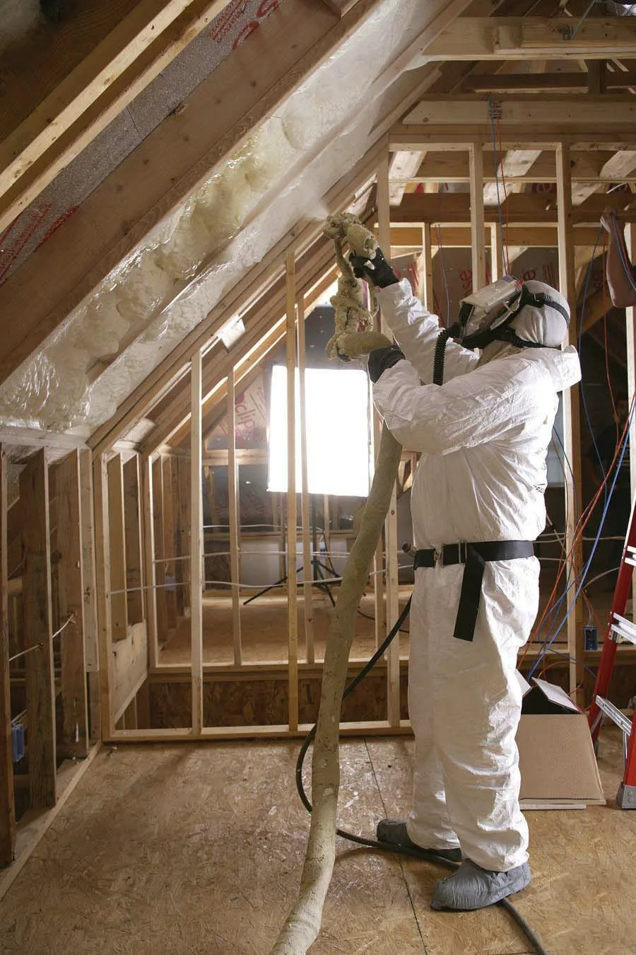 Technician in protective gear spraying foam insulation on the underside of an attic roof during construction.